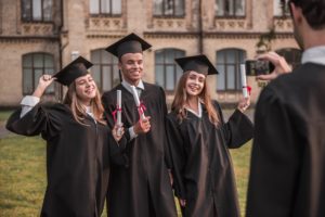Odnoszący sukcesy absolwenci w strojach akademickich trzymają dyplomy, patrzą w aparat i uśmiechają się, stojąc na zewnątrz, facet robi im zdjęcie