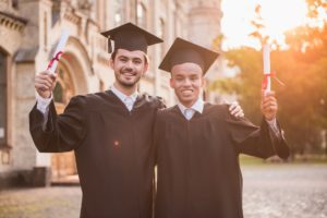 Przystojni absolwenci w akademickich sukienkach trzymają dyplomy, patrzą w kamerę i uśmiechają się, stojąc na zewnątrz